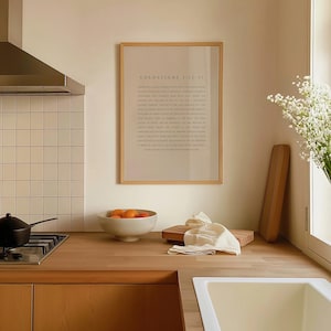 May include: A framed print with the text "Colossians 1:12-17" in a light brown frame hanging on a white wall in a kitchen. The kitchen has a wooden countertop, a stainless steel stovetop, and a white sink.