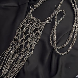 Könnte beinhalten: Eine dunkelgraue, metallisch glänzende Umhängetasche mit geknotetem Netzdesign und langen Fransen am unteren Ende. Die Tasche verfügt über einen passenden geflochtenen Schulterriemen, der mit silberfarbenen Metallringen befestigt ist.