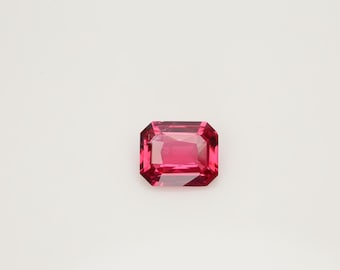 May include: A faceted, rectangular-cut, vibrant pink gemstone. The gem has a symmetrical shape with multiple facets that reflect light, creating a sparkling effect. The gemstone is set against a plain white background.