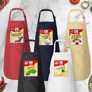 May include: A collection of five aprons in various colors: red, black, navy blue, white, and tan. Each apron features a pocket and a label with the "SG" logo and the name of a spice: Jamaican Jerk Seasoning, Kosher Salt, Chinese Five Spice, Basil, and Ground Ginger.