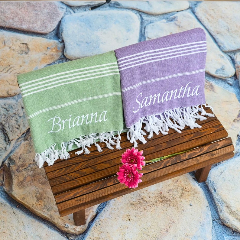 Two Turkish beach towels on a wooden bench with personalized embroidery towel. Custom design with name and log. Both have white stripes and fringed edges, perfect for custom gifts, beach trips, and special occasions.