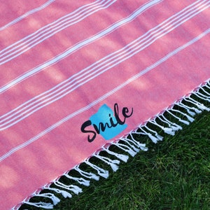 A pink cotton towel with white stripes and tassels, featuring the word Smile in black and blue embroidery. Placed on green grass, it is ideal for personalized beach towels with your logo, pool towels for coworkers, and appreciation gifts.