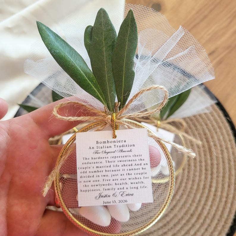 May include: A circular gold-toned metal frame containing white sugared almonds wrapped in white tulle, topped with green olive leaves and a jute twine bow. A square white tag features the text Bomboniera An Italian Tradition, Justin & Erica, June 13, 2026.