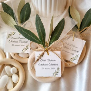 Könnte beinhalten: Nahaufnahme von Taufgeschenken. Jedes Geschenk zeigt ein kleines, rundes, beiges Objekt, das mit Olivenzweigen und einem Etikett verziert ist. Auf den Etiketten steht "Sante Battesimo Cristiano Cavalieri 29 marzo 2026". Die Geschenke sind mit Bindfaden und Tüll gebunden.