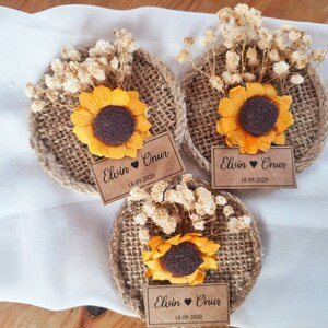May include: Three round burlap and twine ornaments with a dried flower arrangement and a sunflower in the center. Each ornament has a brown paper tag with the text "Elvin &hearts; Onur 18.09.2020".