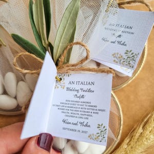 May include: A wedding favor featuring white sugared almonds wrapped in sheer tulle with green olive leaves and a twine bow. A small white card is attached, printed with the text: AN ITALIAN Wedding Tradition Confetti, Kim and Thika, September 19, 2027.