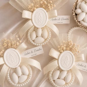 Pode incluir: Três lembranças de casamento com tule branco, fitas de cetim creme e flores secas. Cada lembrança tem um selo de cera oval com as letras A e N, uma borda de pequenas pérolas brancas e amêndoas brancas. Uma etiqueta diz Amber & Demitri 12 MAY 2025.