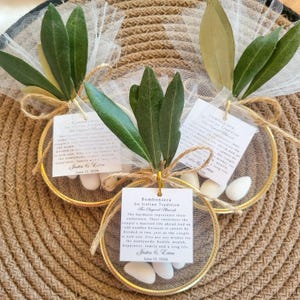 May include: Three circular gold-toned metal hoops containing white tulle fabric and five white almond candies. Each hoop is decorated with green olive leaves, a jute twine bow, and a square white tag printed with the text Bomboniera An Italian Tradition Five Sugared Almonds, Justin & Erica, June 13, 2026.