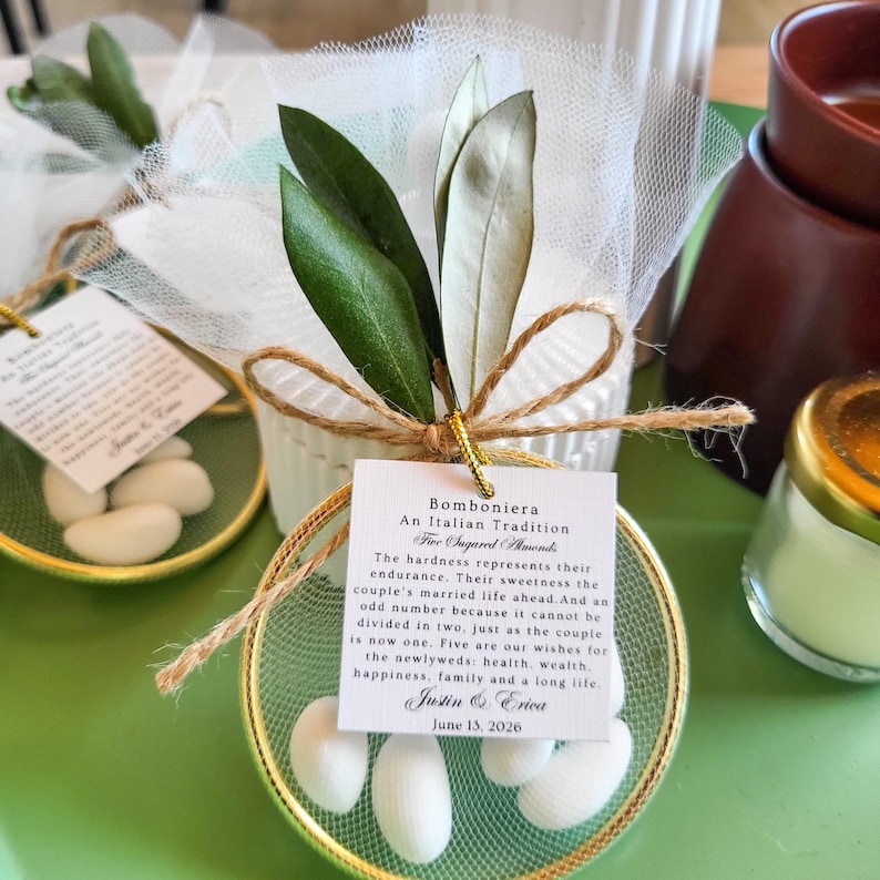 May include: A round gold-rimmed favor container filled with five white sugared almonds, wrapped in white tulle, and topped with two green olive leaves and a jute twine bow. A square white tag features the text Bomboniera An Italian Tradition.