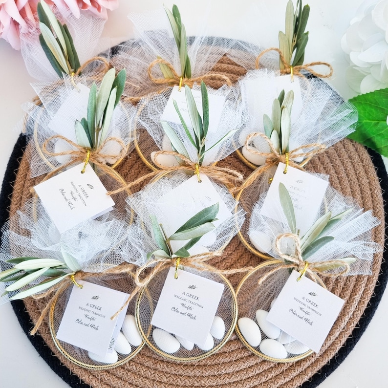 May include: A collection of circular gold-toned metal hoops containing white tulle fabric, filled with white sugared almonds. Each hoop is decorated with a green olive branch, natural twine, and a square white tag labeled A GREEK WEDDING TRADITION.