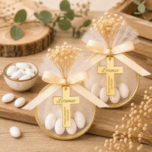 May include: Two circular gold-rimmed containers filled with white almond candies, wrapped in cream-colored tulle, and tied with a matching ribbon. A gold cross charm hangs from each ribbon, engraved with the name Lorenzo and Prima Comunione 24.05.2023. Dried baby's breath flowers are tucked into the top of each tulle bundle.