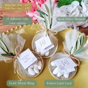 May include: Three wedding favors featuring a gold metal ring, white tulle, a real olive branch, and a folded label card. The ring measures 7cm or 2.7 inches, and the tulle measures 12cm or 4.7 inches. Each favor contains white sugared almonds.