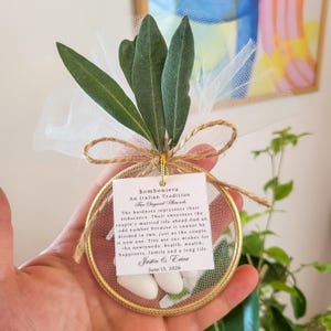 May include: A circular gold-rimmed favor containing white sugared almonds wrapped in white tulle, topped with green olive leaves and a jute twine bow. A square white tag reads: Bomboniera An Italian Tradition, Five Sugared Almonds, Justin & Erica, June 13, 2026.