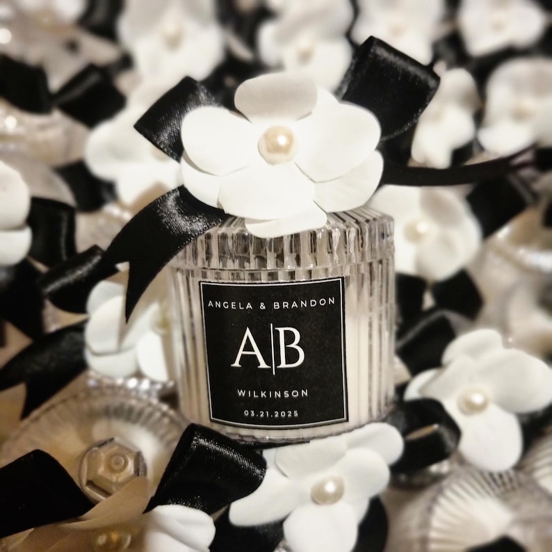 Pu&ograve; includere: Un barattolo per candela in vetro trasparente a coste sormontato da un fiore bianco e un nastro di raso nero. L'etichetta riporta il testo Angela & Brandon, le iniziali AB, il nome Wilkinson e la data 03.21.2025 stampati in bianco su fondo nero.