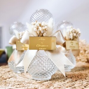 May include: Clear glass bottle favors with a gold ribbon and a gold tag that says "John & Christina 10.07.2023". The bottles are decorated with dried white flowers.
