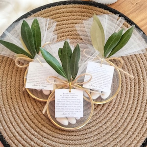 May include: Three circular gold-toned metal hoops decorated with white tulle fabric, green olive leaves, and jute twine bows. Each hoop contains white sugar-coated almonds and a square white paper tag with printed text detailing an Italian tradition.