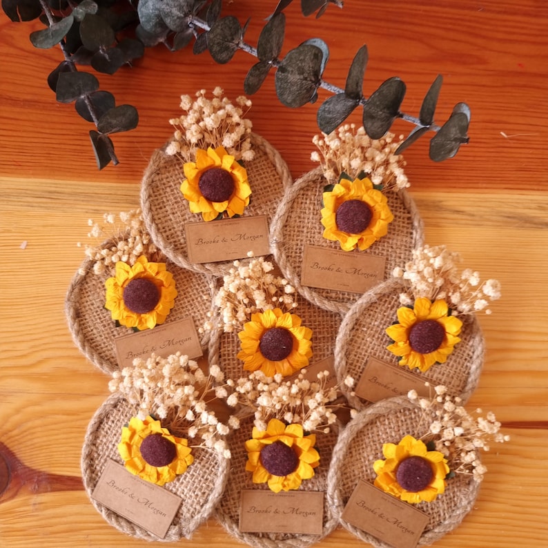 May include: Nine round burlap coasters with a sunflower and dried white flowers on top. Each coaster has a small brown tag with the text "Brenda & Morgan" printed on it.