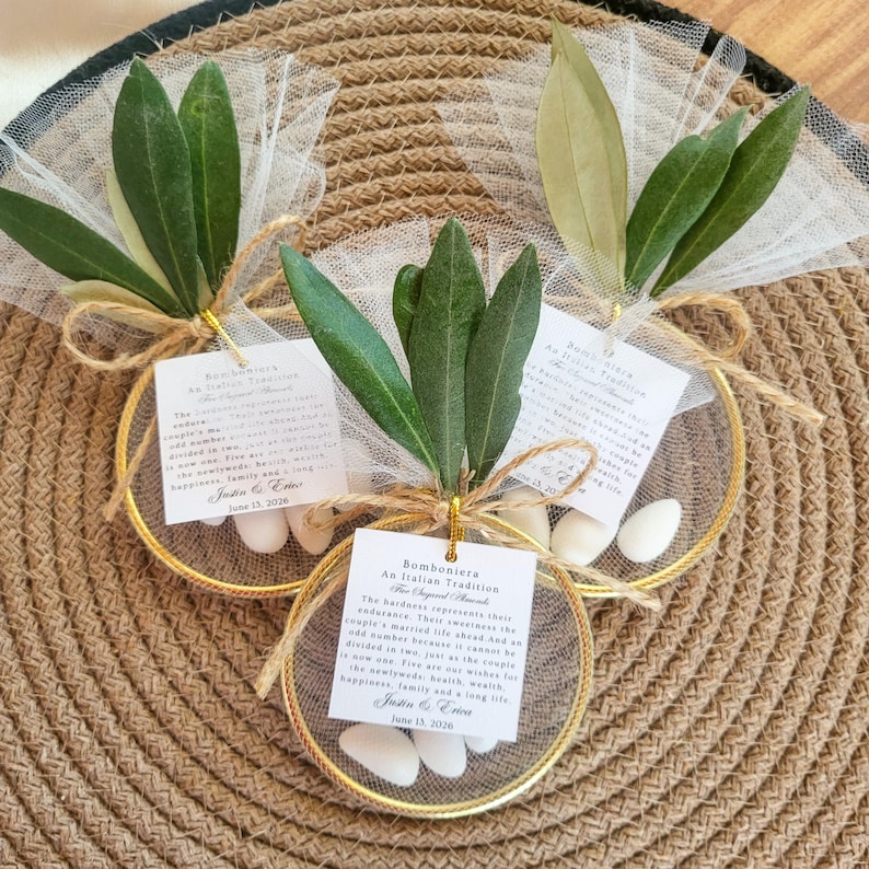 May include: Three circular gold-toned metal hoops decorated with white tulle, green olive leaves, and white almond candies. Each hoop features a square white tag with black text reading Bomboniera An Italian Tradition, Justin & Erica, June 13, 2026.
