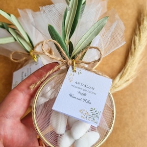 May include: A round gold metal hoop filled with white tulle and white almond candies, tied with twine and a green olive branch. A square white tag is attached, printed with the text AN ITALIAN WEDDING TRADITION Confetti Kim and Thika.