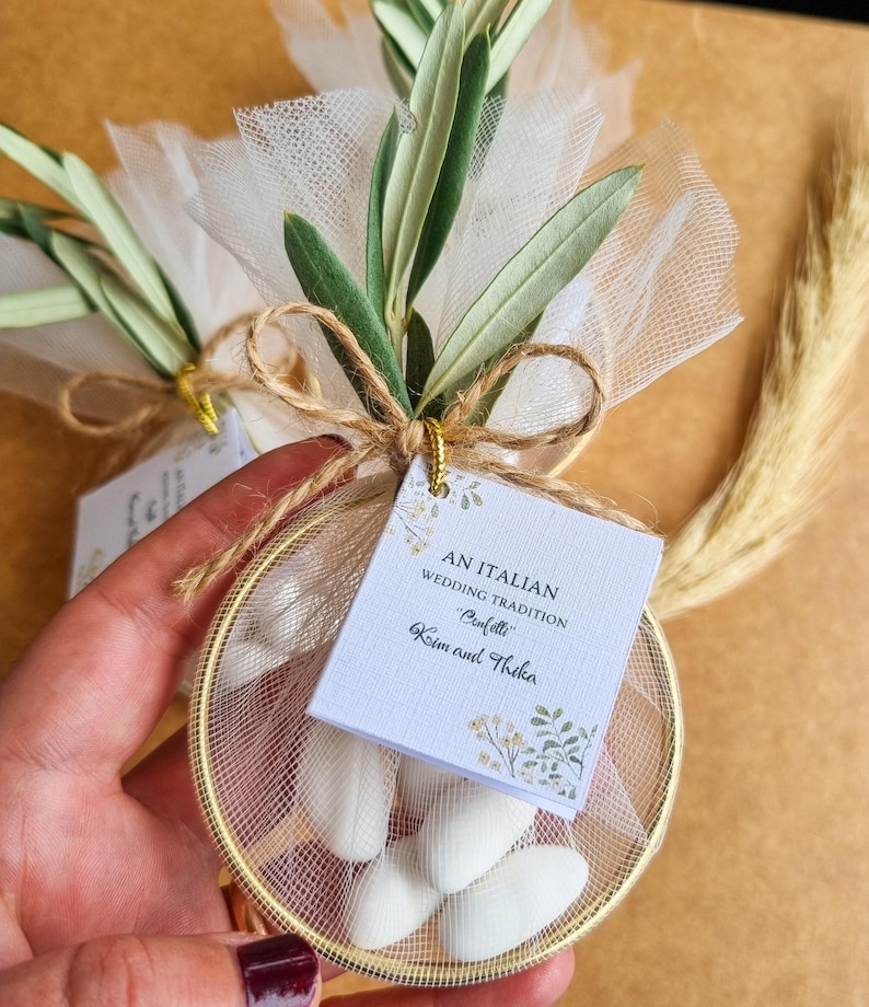 May include: A round gold metal hoop filled with white tulle and white almond candies, tied with twine and a green olive branch. A square white tag is attached, printed with the text AN ITALIAN WEDDING TRADITION Confetti Kim and Thika.