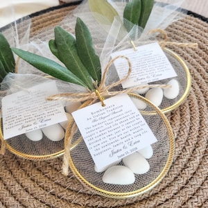 May include: A circular gold-toned metal hoop containing white sugared almonds wrapped in sheer white tulle. Each favor features a green olive branch sprig, a jute twine bow, and a square white card printed with the text Bomboniera An Italian Tradition.