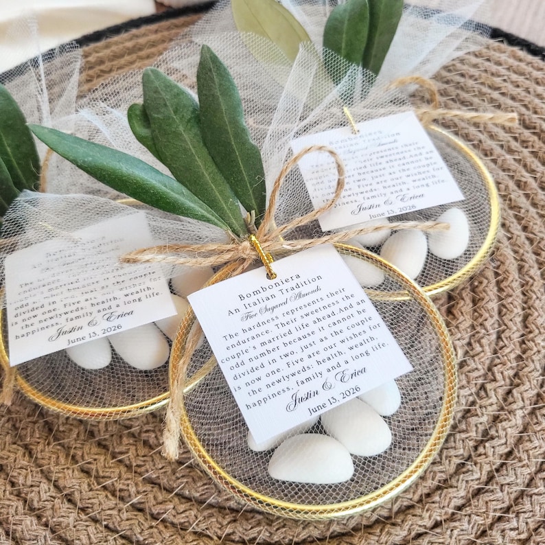 May include: A circular gold-toned metal hoop containing white sugared almonds wrapped in sheer white tulle. Each favor features a green olive branch sprig, a jute twine bow, and a square white card printed with the text Bomboniera An Italian Tradition.
