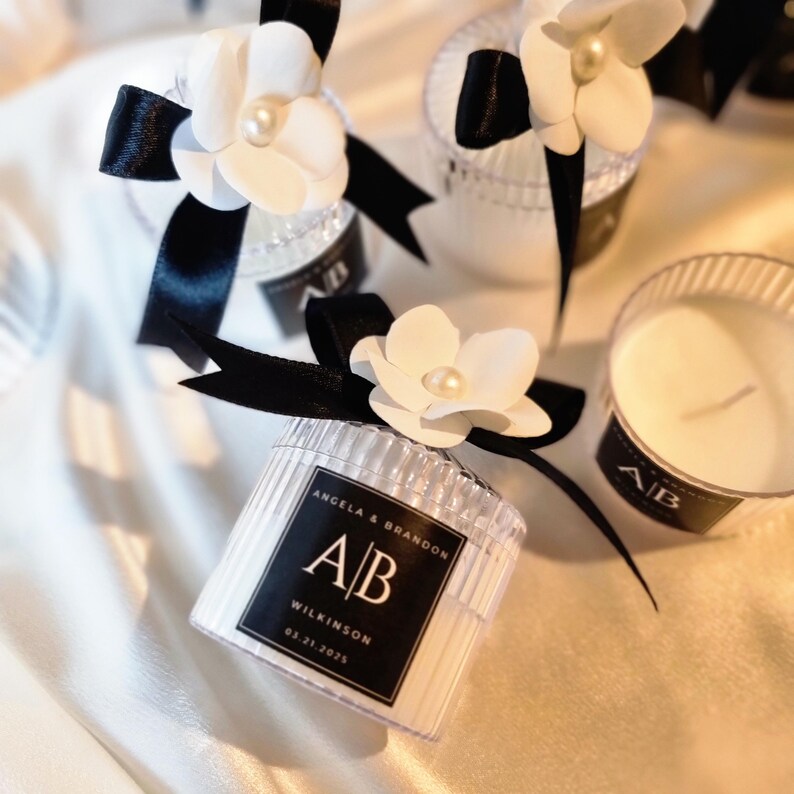 Pu&ograve; includere: Candele in barattoli di vetro trasparente a coste con cera bianca, decorate con un nastro di raso nero e un fiore bianco con una perla centrale. Un'etichetta nera riporta il testo Angela & Brandon, A|B, Wilkinson, 03.21.2025.