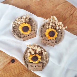 May include: Three round burlap favors with a sunflower and dried white flowers. Each favor has a brown paper tag with the text "Elvin &hearts; Onur 18.09.2020"