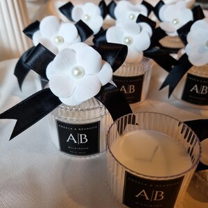 Pu&ograve; includere: Un gruppo di piccole candele in barattoli di vetro trasparente a coste, ognuna con un'etichetta quadrata nera con il testo "ANGELA & BRANDON", "A|B", "WILKINSON" e "04.21.2025". Ogni candela &egrave; decorata con un fiore in tessuto bianco e un fiocco in raso nero.