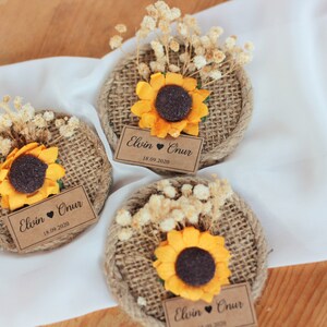 May include: Three round burlap favors with a sunflower and dried flowers on top. Each favor has a brown paper tag with the text "Elvin &hearts; Onur 18.09.2020"