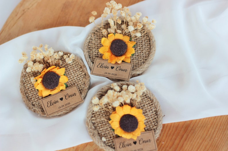 May include: Three round burlap favors with a sunflower and dried flowers on top. Each favor has a brown paper tag with the text "Elvin &hearts; Onur 18.09.2020"