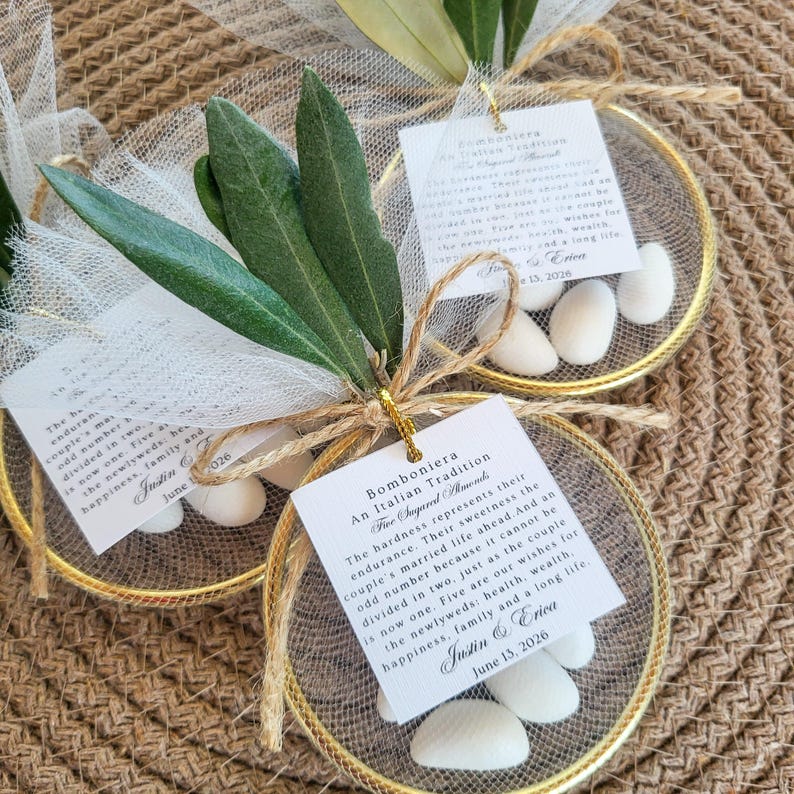 May include: Two circular gold-toned metal frames filled with white tulle netting, each containing five white sugared almonds. Each favor is tied with twine and features a white card printed with the text Bomboniera An Italian Tradition.