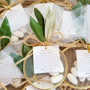 May include: A collection of wedding favors featuring white sugared almonds inside a gold circular frame wrapped in white tulle. Each favor includes a green olive branch, a jute twine bow, and a square white tag with printed text about the tradition.