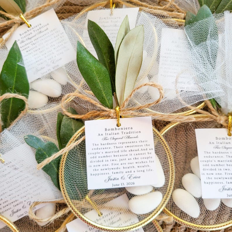 May include: A collection of wedding favors featuring white sugared almonds inside a gold circular frame wrapped in white tulle. Each favor includes a green olive branch, a jute twine bow, and a square white tag with printed text about the tradition.