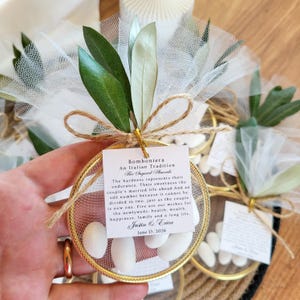 May include: A circular gold-rimmed favor containing white sugared almonds wrapped in white tulle, adorned with green olive leaves and a jute twine bow. A square white tag features the text Bomboniera An Italian Tradition, Justin & Erica, June 13, 2026.