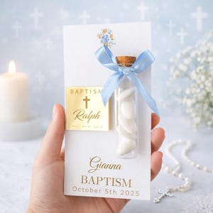Puede incluir: Un recuerdo de bautizo con un vial de vidrio transparente lleno de caramelos blancos, atado con una cinta azul claro. El vial está unido a una tarjeta blanca con detalles dorados y el texto "Bautizo" y un nombre.