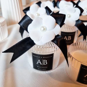 Pu&ograve; includere: Un barattolo per candela in vetro trasparente a coste sormontato da un fiore bianco e un nastro di raso nero. Un'etichetta rettangolare nera riporta il testo Angela & Brandon, le iniziali AB, il nome Wilkinson e la data 21.03.2025 in bianco.