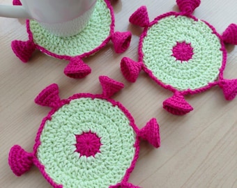 May include: Three handmade crochet coasters. Each coaster is circular with a light green center, a bright pink border, and matching pink decorative elements. A white mug rests on one coaster, with the other two coasters nearby on a light wood surface.