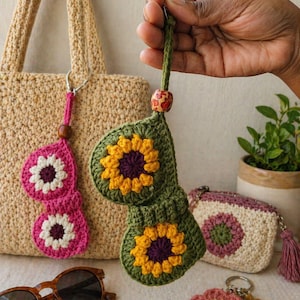 Könnte beinhalten: Eine Sammlung handgefertigter Häkelartikel, darunter ein grüner Sonnenblumen-Schlüsselanhänger, eine rosa-weiße Sonnenbrillenhalterung, eine kleine Geldbörse und ein Blumen-Schlüsselanhänger. Eine gewebte Tragetasche und Sonnenbrille sind ebenfalls zu sehen.