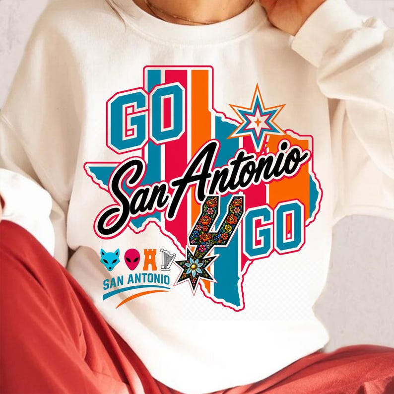 Puede incluir: Una sudadera blanca de cuello redondo con un gr&aacute;fico del estado de Texas en rayas verde azulado y naranja. El texto dice GO San Antonio GO. Incluye un icono de espuela con estampado floral, una estrella y peque&ntilde;os iconos de un zorro, alien, castillo y arpa.