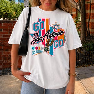 Puede incluir: Una camiseta blanca de manga corta con un gr&aacute;fico colorido del estado de Texas y el texto GO San Antonio GO. El dise&ntilde;o incluye una estrella, una espuela con estampado floral y peque&ntilde;os iconos de un zorro, alien&iacute;gena, torre y arpa.
