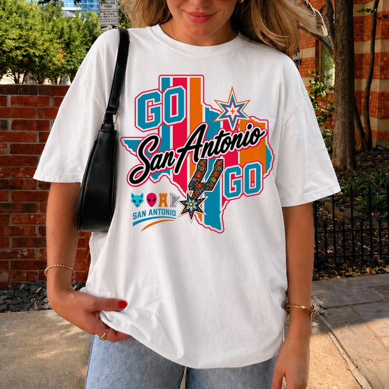 Puede incluir: Una camiseta blanca de manga corta con un gr&aacute;fico colorido del estado de Texas y el texto GO San Antonio GO. El dise&ntilde;o incluye una estrella, una espuela con estampado floral y peque&ntilde;os iconos de un zorro, alien&iacute;gena, torre y arpa.