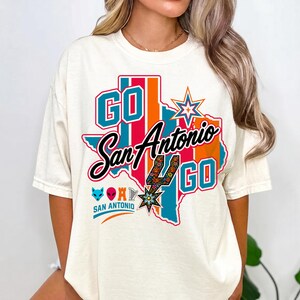 Puede incluir: Una camiseta de manga corta color hueso con un gr&aacute;fico del estado de Texas en rayas verde azulado y naranja. El texto dice GO San Antonio GO. El dise&ntilde;o incluye una espuela estilizada, una estrella e iconos de un zorro, alien y arpa.