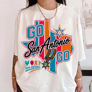 Puede incluir: Una camiseta color hueso con un gr&aacute;fico del estado de Texas en rayas azules, naranjas y rosas. El texto dice GO San Antonio GO. Incluye un logotipo de espuela estilizado, una estrella, un icono de zorro, una cabeza alien&iacute;gena, una torre y una arpa.