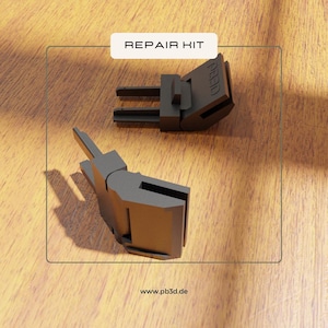Puede incluir: Dos componentes de kit de reparación de plástico gris oscuro se muestran sobre una superficie de madera. El texto "REPAIR KIT" está en un rectángulo blanco en la parte superior. La dirección del sitio web "www.pb3d.de" está en la parte inferior.
