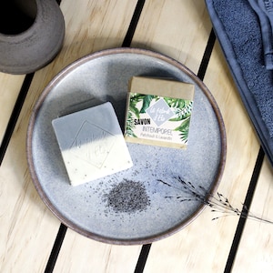 Peut inclure: Deux savons sur une assiette en céramique. L'un est gris clair et blanc avec le texte "La Fabrik d'Elo". L'autre est un petit savon emballé avec le texte "Savon Intemporel Patchouli & Lavandin". Un petit tas de graines est à côté du savon.