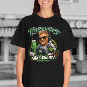 May include: A black t-shirt featuring a graphic of a cartoon turtle character wearing sunglasses and headphones, holding a green tumbler and sitting at a laptop. The text above and below the character reads TURTLEBOY WAS RIGHT!