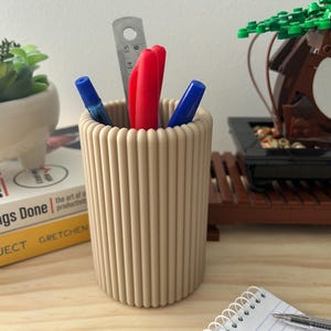 May include: A beige, ribbed pen holder filled with pens, markers, and a ruler. The holder sits on a wooden desk next to a notepad and a small bonsai tree. The pens are red and blue.
