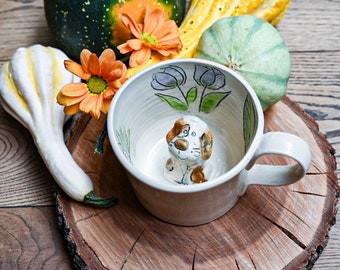 Puede incluir: Una taza de cerámica blanca con una ilustración de perro marrón y blanco en su interior. La taza tiene un diseño floral en el exterior y un asa en el lado derecho. La taza está sentada sobre una superficie de madera con calabazas y flores alrededor.