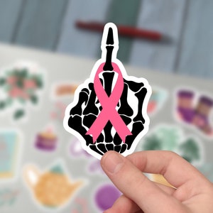 May include: A black and white sticker featuring a skeletal hand giving the middle finger, with a pink ribbon tied around the middle finger. The sticker is held in a person's hand, with a blurred background of other stickers and a laptop.